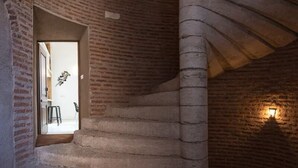 Un escalier en colimaçon dans un intérieur aux murs de briques, avec une petite fenêtre et un luminaire fixé au mur.