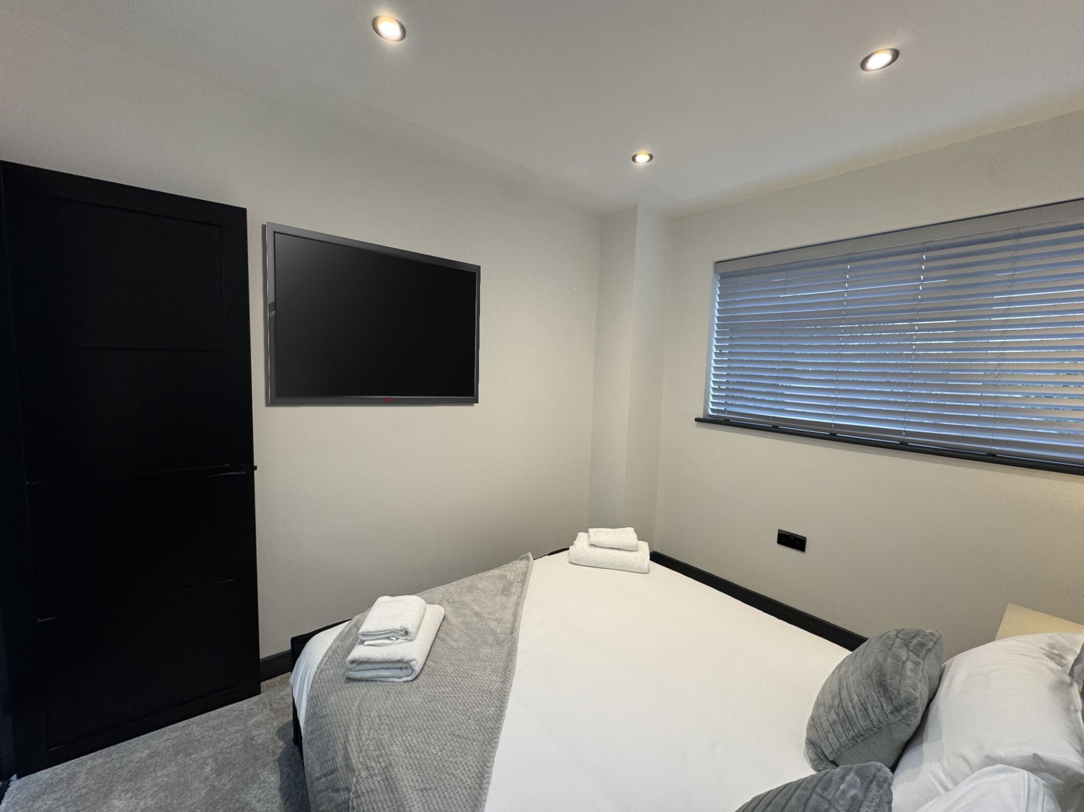 A modern hotel room with a bed, a flat-screen TV mounted on the wall, and a window with blinds.