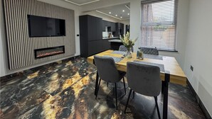 A modern dining area with a wooden table, grey chairs, a fireplace, and a kitchen in the background. - Miles House (Boston)