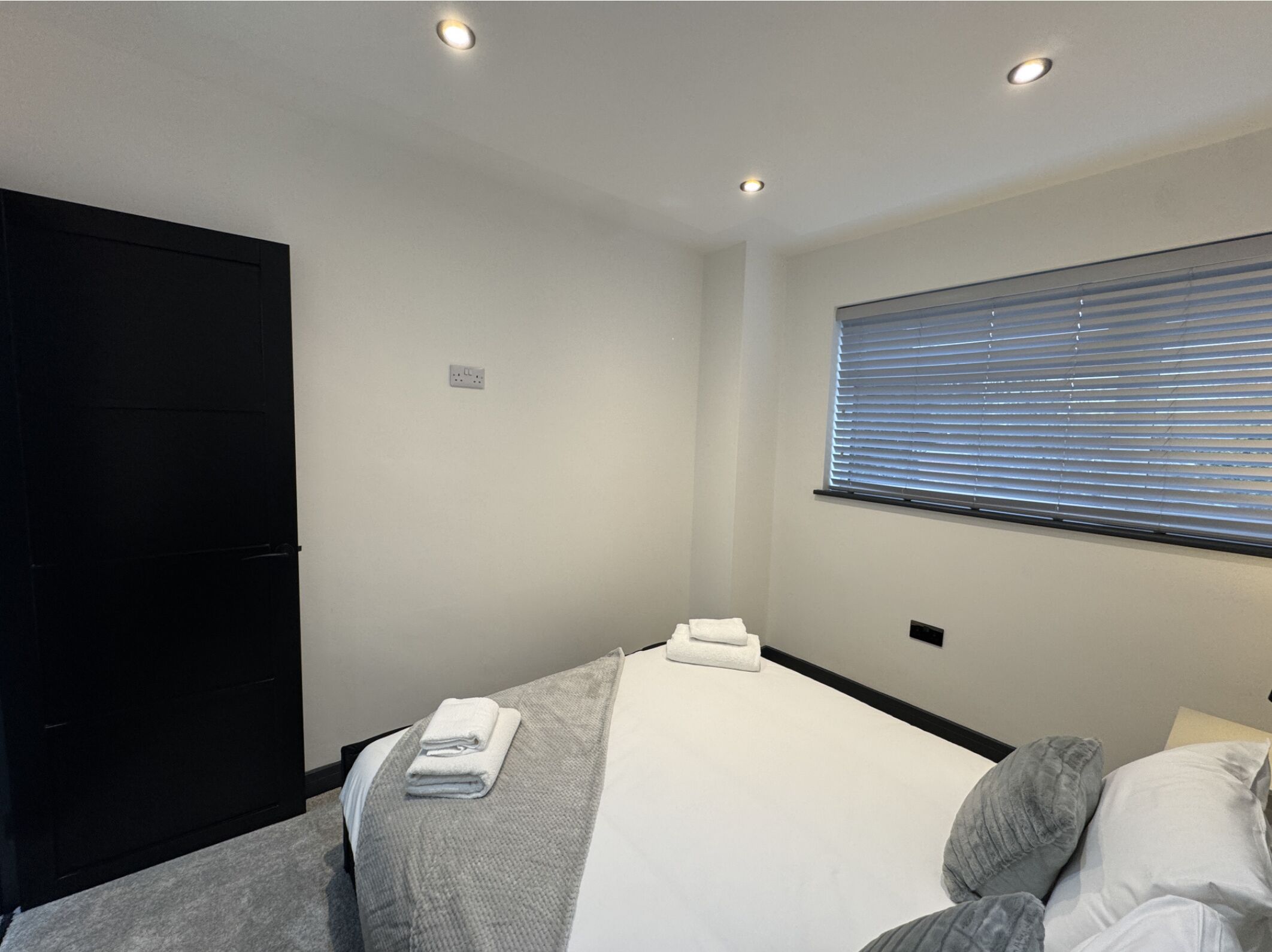 Ein Schlafzimmer mit einem Bett, einem Fenster mit Jalousien, einem schwarzen Kleiderschrank und Einbauleuchten.