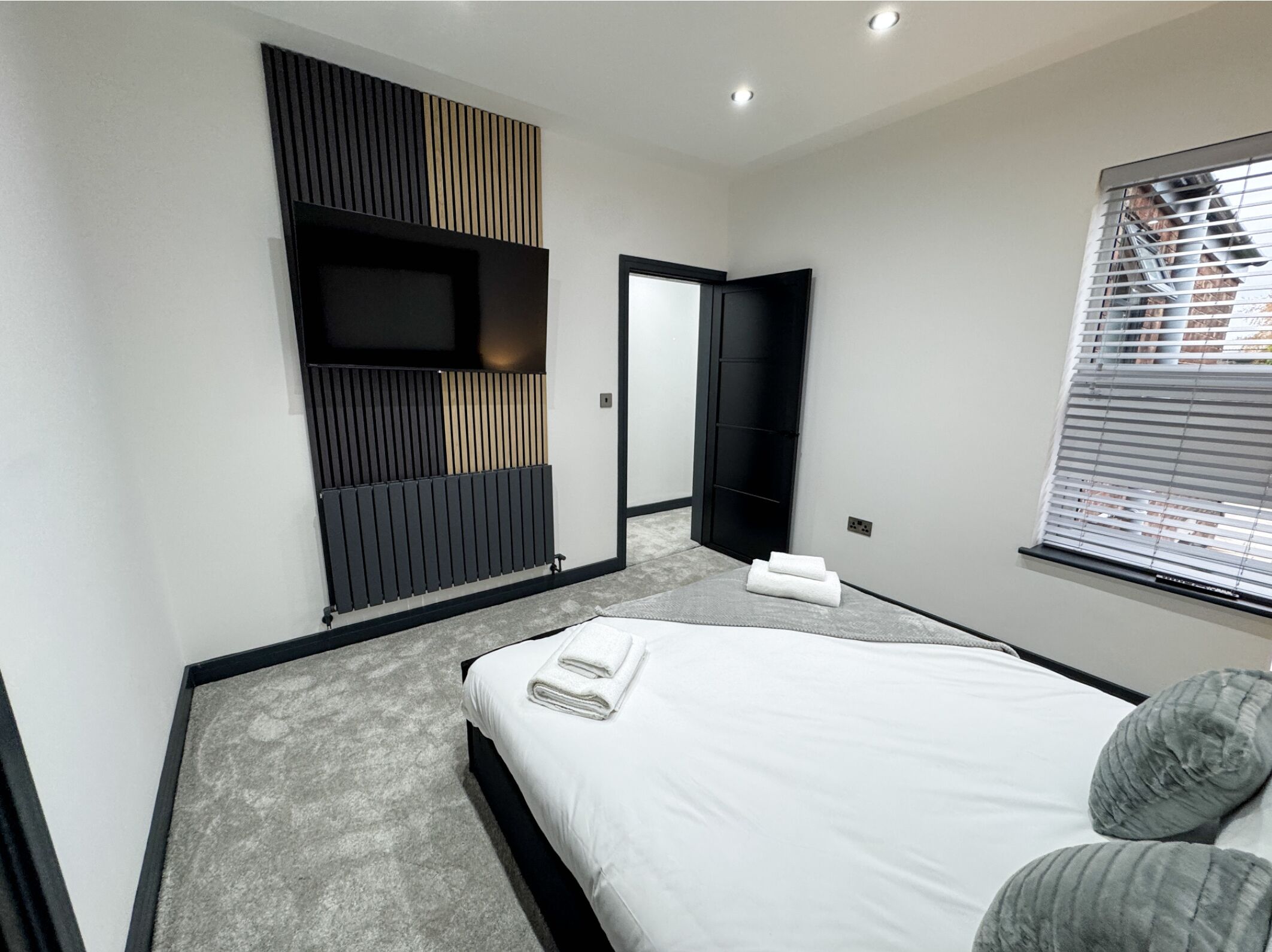 A modern bedroom with a bed, a built-in TV, a large mirror, and a window with blinds.