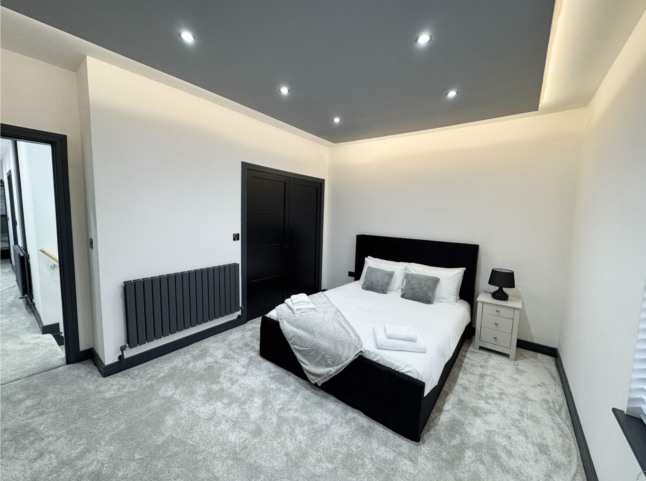 Un dormitorio moderno con una cama negra, paredes blancas, un radiador, una mesita de noche con una lámpara y una puerta.