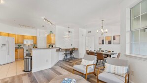A modern kitchen with wooden cabinets, a central island, and pendant lighting. There is a dining area with a table and chairs, and a living area with a sofa and a coffee table.