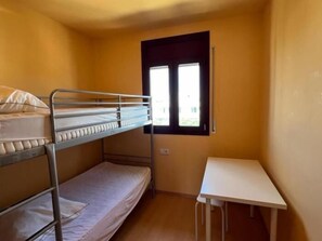 A small, single-room with a bunk bed, a small table, and a window.