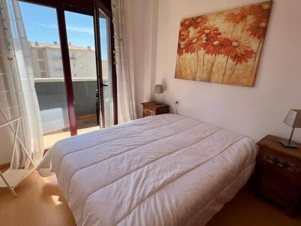 A bedroom with a large bed, a painting of flowers on the wall, a nightstand, and a view of buildings through a sliding door.
