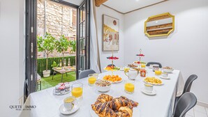 A breakfast table set with croissants, fruit, and orange juice in a bright room with a view of a garden and stone wall. - Renovated House: Peace & Comfort 20 min from Monaco (Peille)