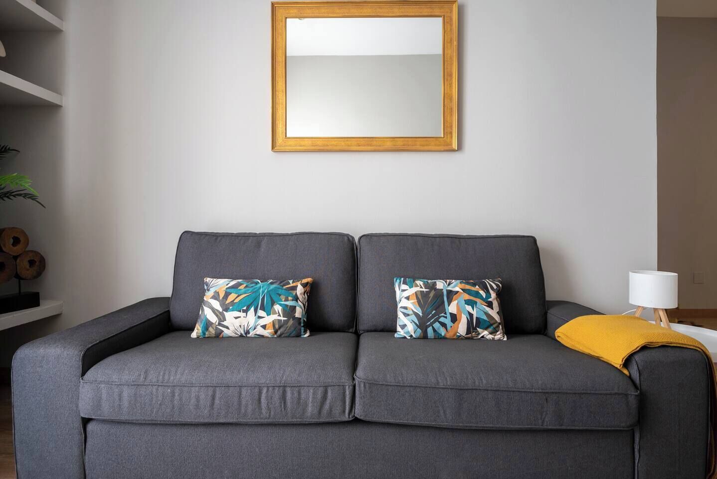 A grey sofa with two patterned cushions, a white lamp on a wooden stand, and a mirror on the wall.