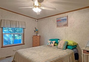Relax in this serene bedroom with a large bed, ceiling fan, and a window offering a view of the surrounding trees and fence.