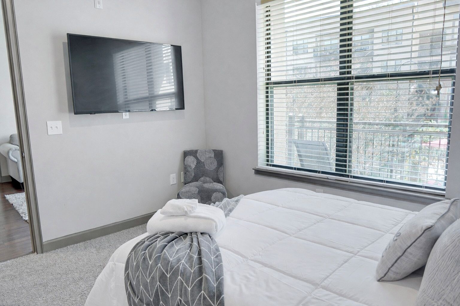 A bedroom with a bed, a TV mounted on the wall, a window with blinds, and a chair with a patterned cushion.