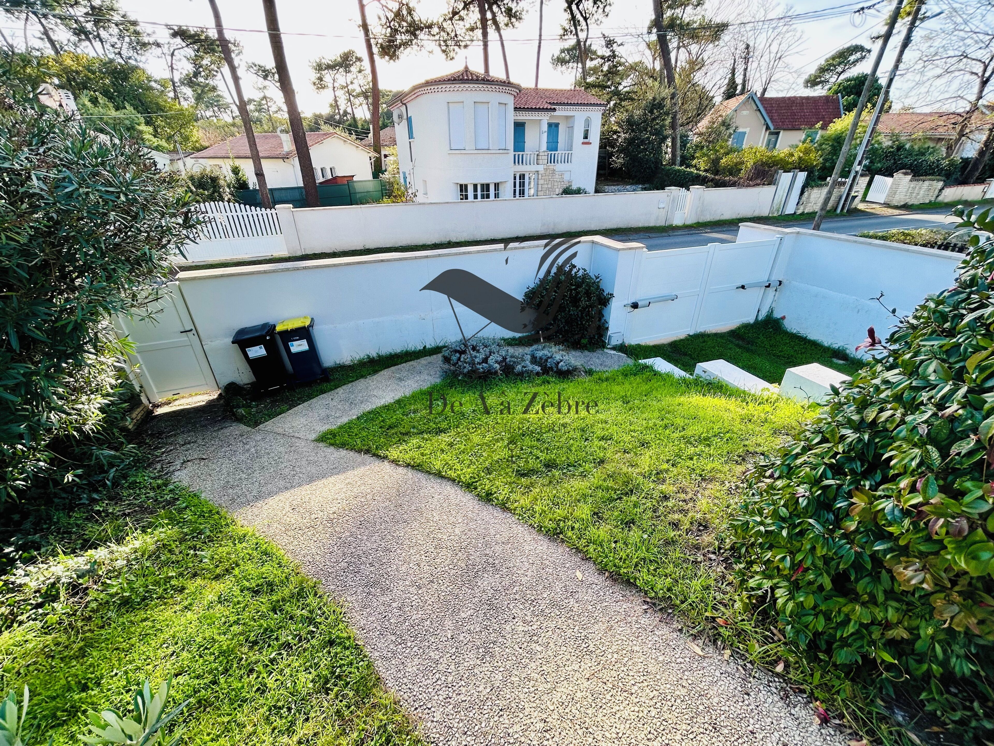 Zona residencial con un sendero pavimentado, una pared blanca y un pequeño jardín con césped y arbustos.