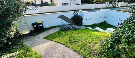 Zona residencial con un sendero pavimentado, una pared blanca y un pequeño jardín con césped y arbustos.