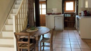 A rustic kitchen with a wooden table and chairs, a hanging light fixture, and a staircase leading to another room.