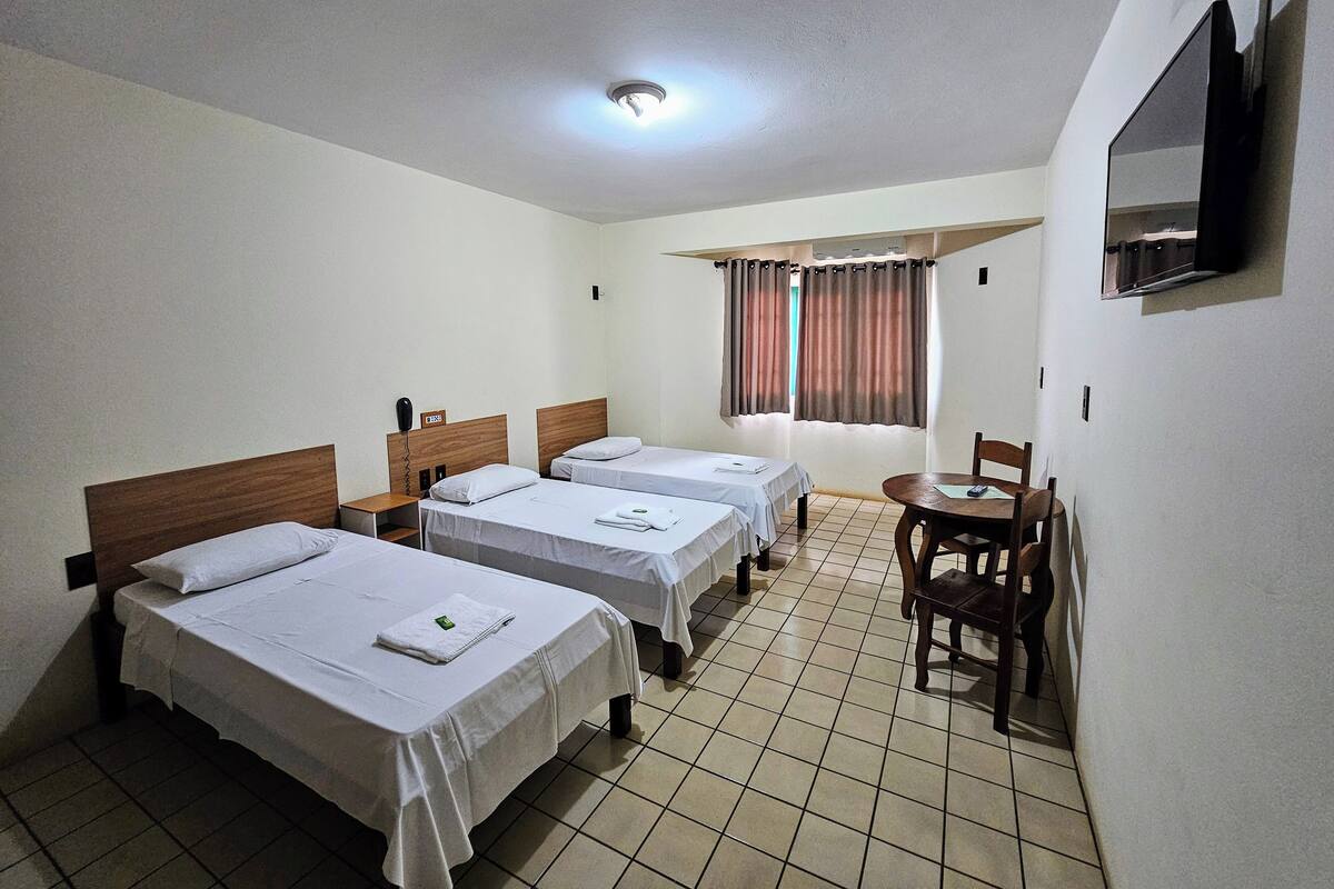 Una habitación de hotel con tres camas individuales, una mesita con sillas, un televisor en la pared y una ventana con cortinas.