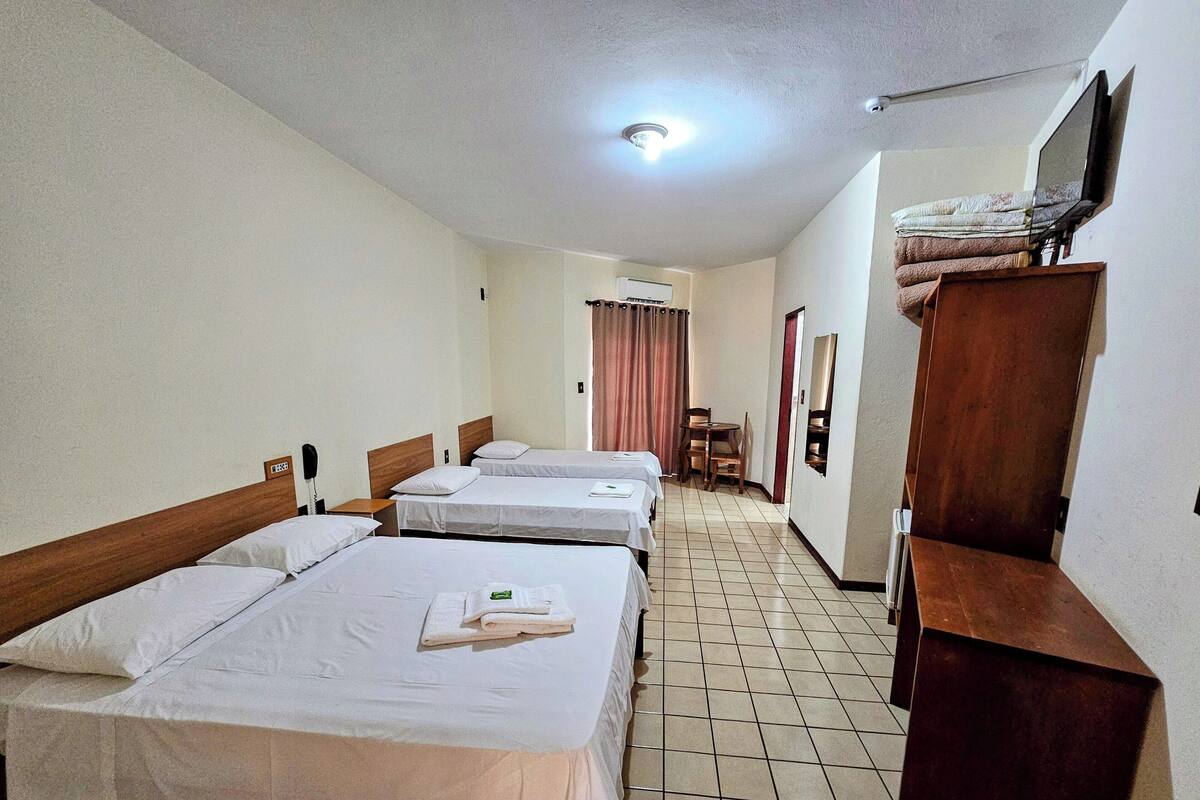 Habitación de hotel con tres camas individuales, un armario de madera, un escritorio y un televisor en la pared.