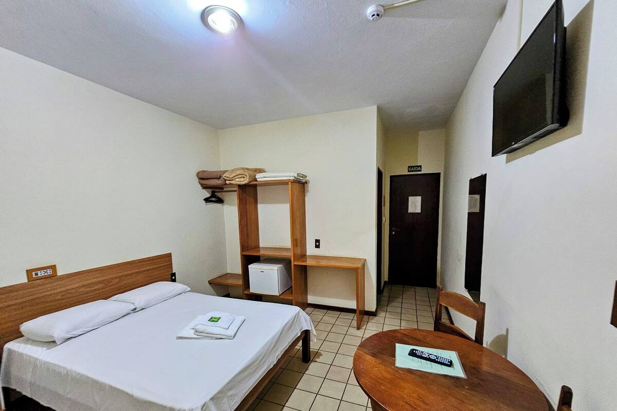 Habitación de hotel con cama, una mesita, una silla, repisas con toallas, un televisor y una puerta con la leyenda 'SAÍDA'.