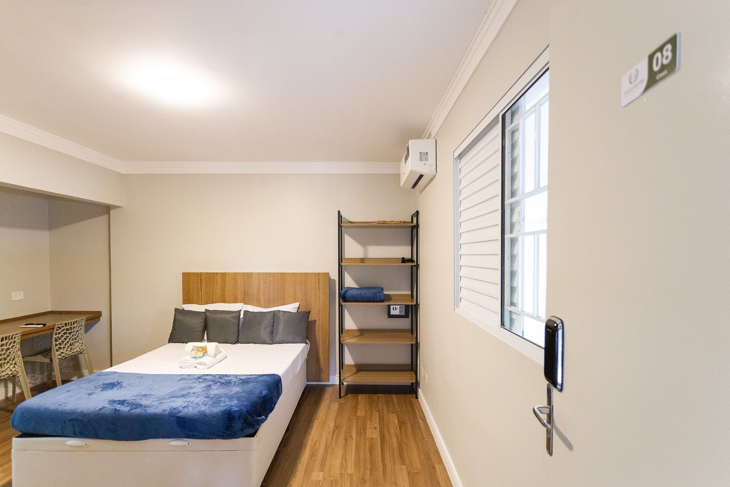 Ein Hotelzimmer mit Bett, Schreibtisch, Regaleinheit und Fenster mit Jalousien.