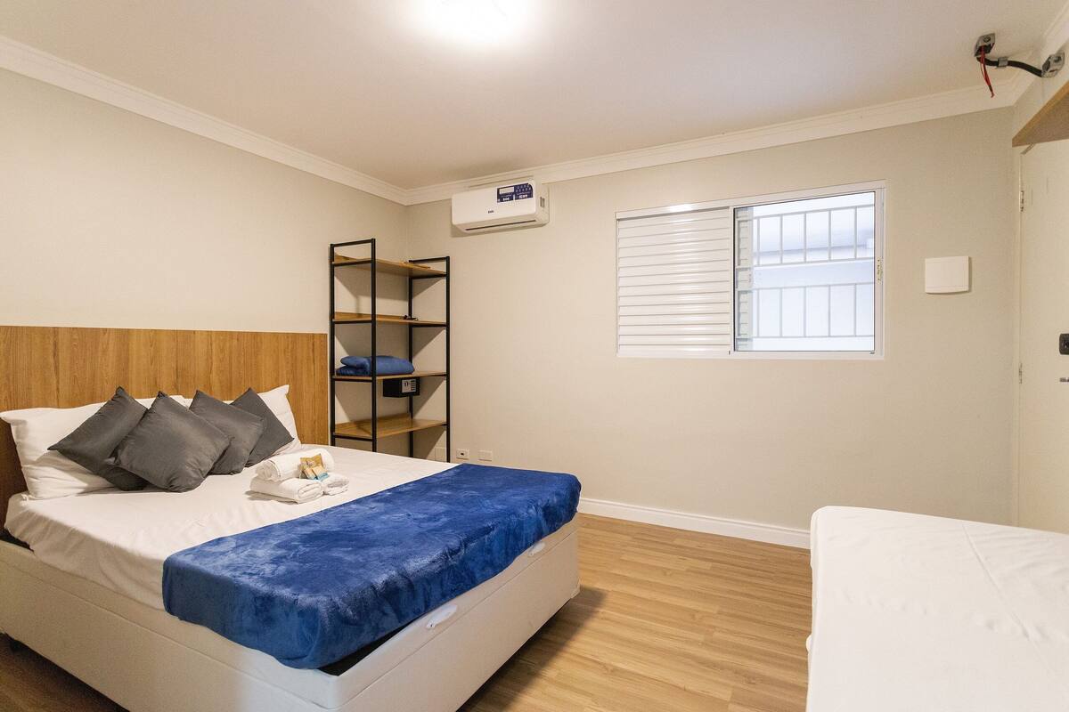 Hotelkamer met een bed, een blauwe deken, een houten hoofdbord, een planken kast en een raam met zonwering.