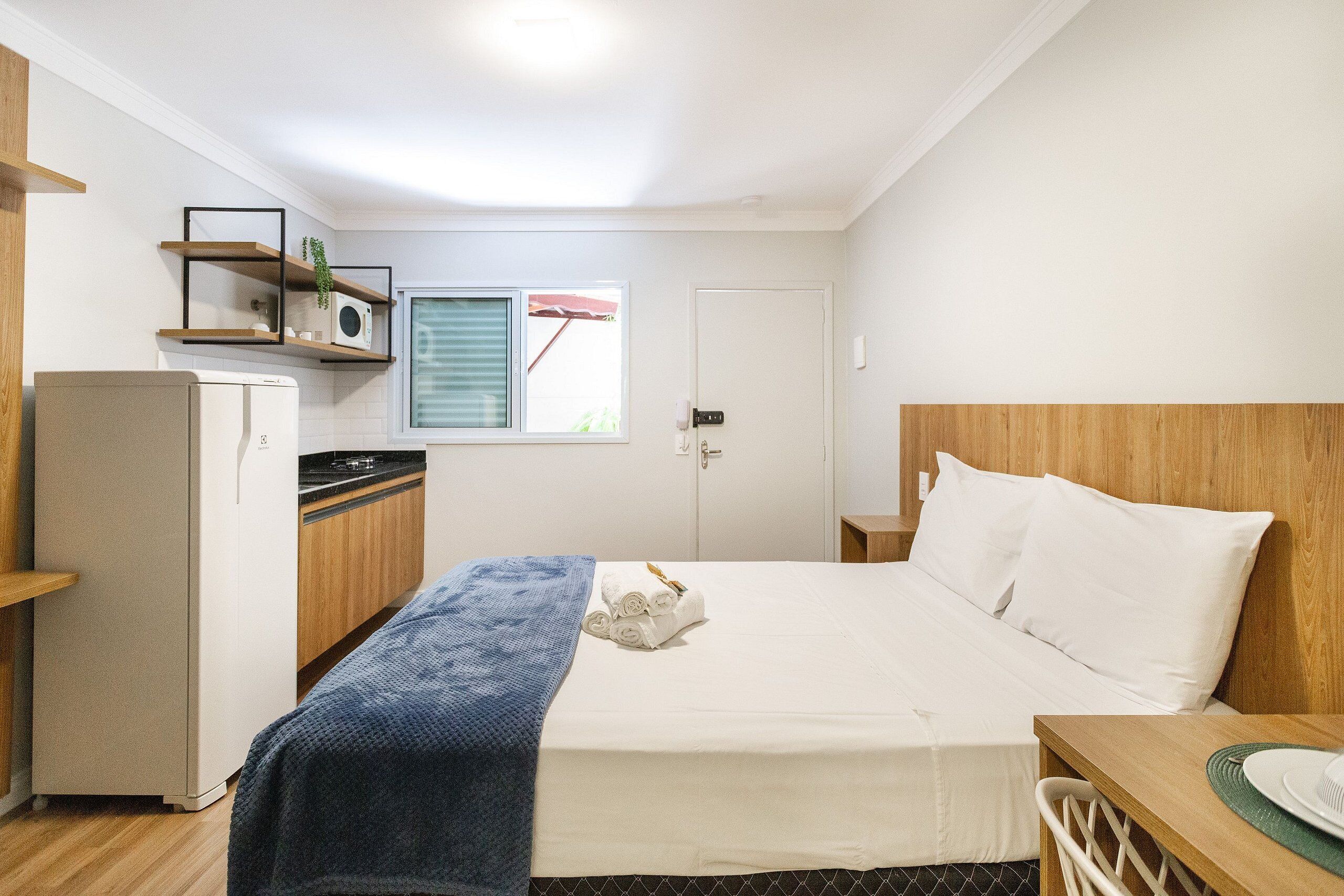 Een compacte hotelkamer met een bed, een kleine kitchenette, een houten hoofdbord en een raam met zonwering.