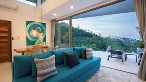 A modern living room with a teal sofa, a dining area, and a large window offering a view of greenery. - ZOG Villas (Koh Samui)