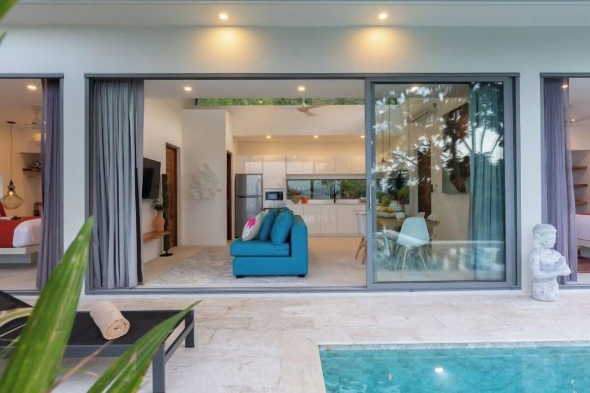 Un espacio moderno de estar con un sofá azul, una piscina y un comedor visibles a través de puertas de cristal.