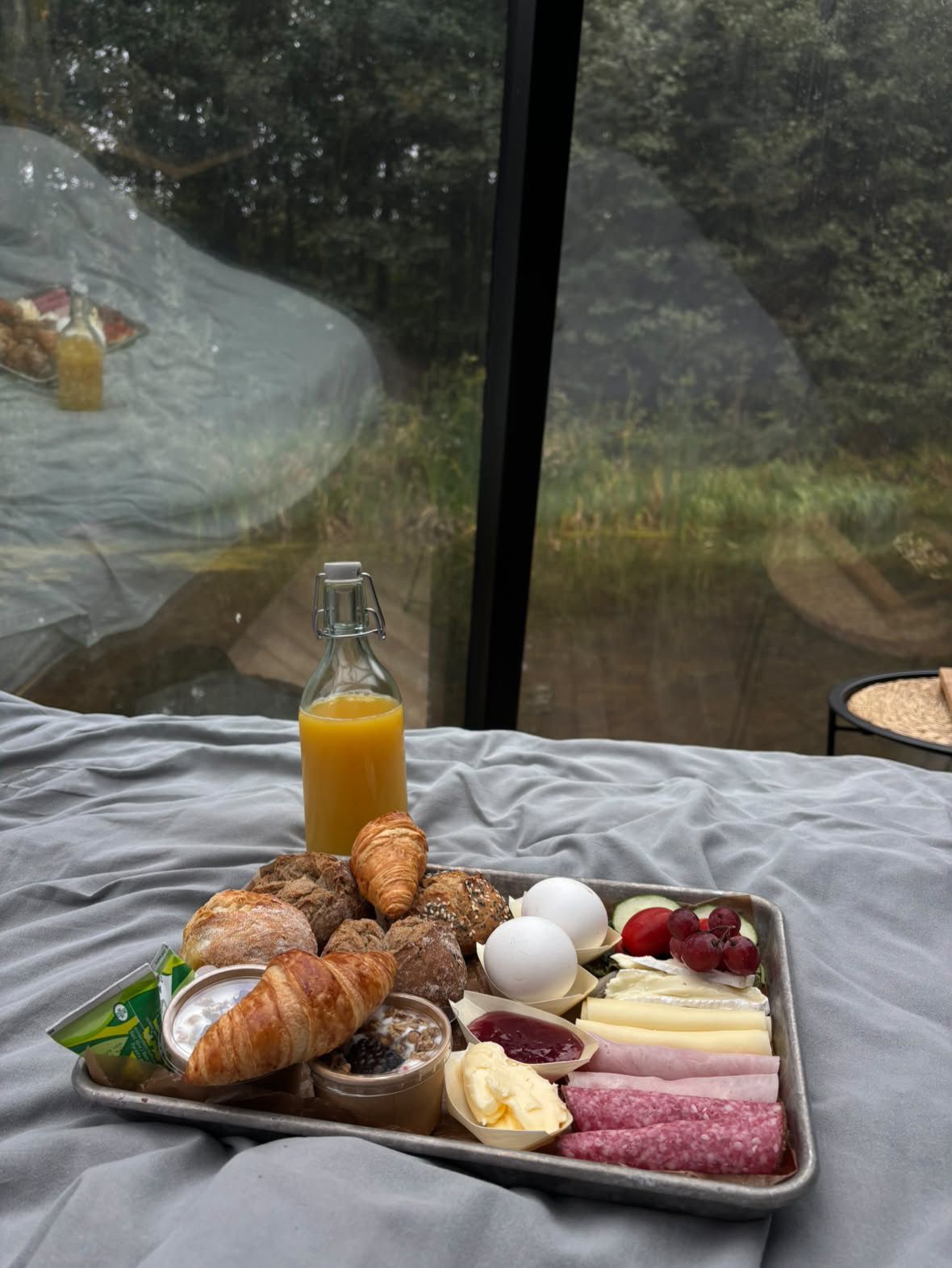 A breakfast tray with croissants, eggs, cheese, ham, and a bottle of orange juice on a bed with a view of trees through a window.
