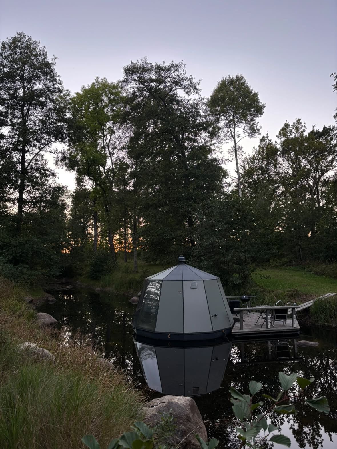 A hexagonal gazebo with a roof, situated by a calm body of water, surrounded by lush greenery and trees.