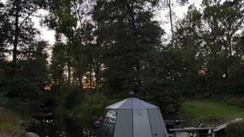 A hexagonal gazebo with a roof, situated by a calm body of water, surrounded by lush greenery and trees.