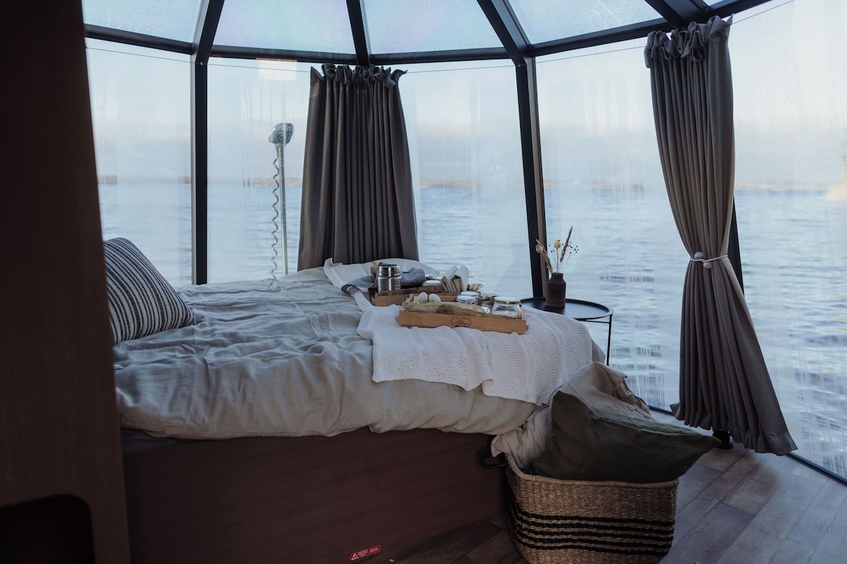 A cozy room with a bed, a small table with a tray, and a view of the water through large windows.