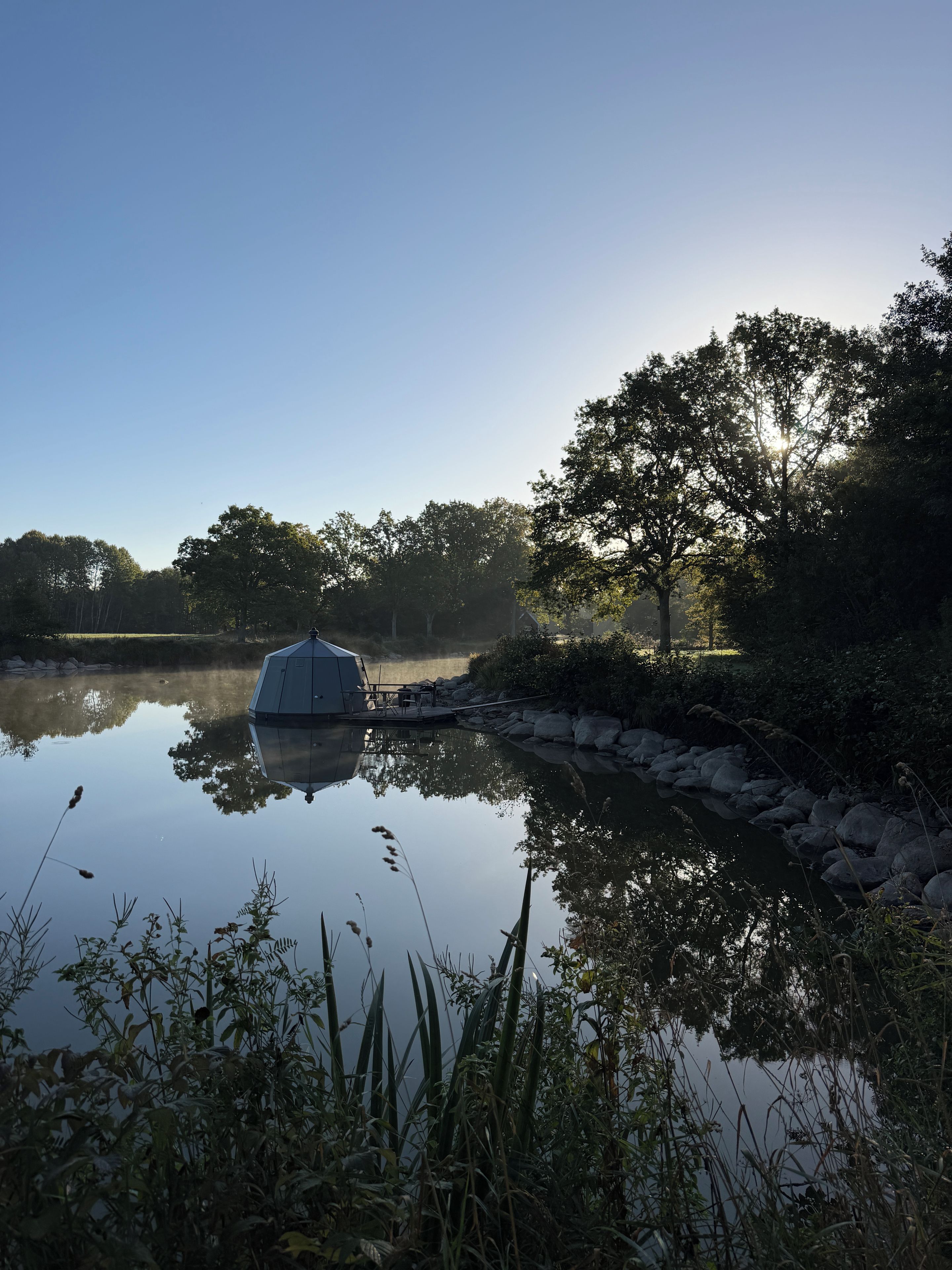 Un paisatge tranquil al costat d'un llac, amb un cobert, aigua calma i arbres al voltant.