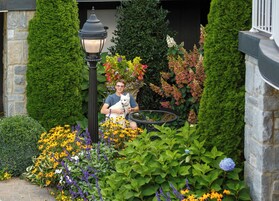 Una persona sosteniendo un perro en un jardín con diversas plantas y una farola.