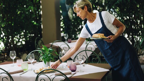 A person serving food at an outdoor table with a potted plant, wine glasses, and a menu. - CONTRA' CONTARINI (Valbrenta)