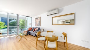 A modern living room with a glass dining table, wooden chairs, a grey sofa, and a large window offering a view of greenery and buildings. - Elegant & Cosy Haven in Lively Kingston Foreshore (Kingston)