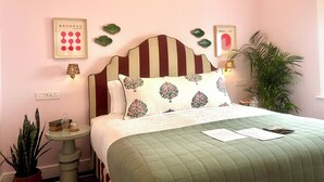 A bedroom with a striped headboard, a green quilted bedspread, and decorative pillows with floral embroidery. There are framed artworks on the wall, a small table with a lamp, and a potted plant beside the bed. - Petit Pineapple House Villa & Guesthouse (Cape Town)