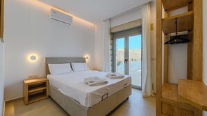A modern hotel room with a large bed, a wooden nightstand, a built-in desk, and a sliding glass door leading to a balcony.