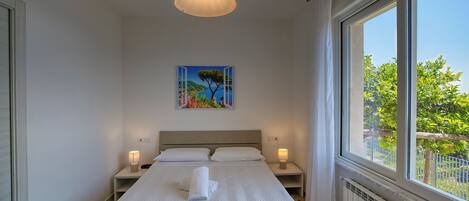 A hotel room with a bed, bedside lamps, a painting, and a large window with a view of greenery.