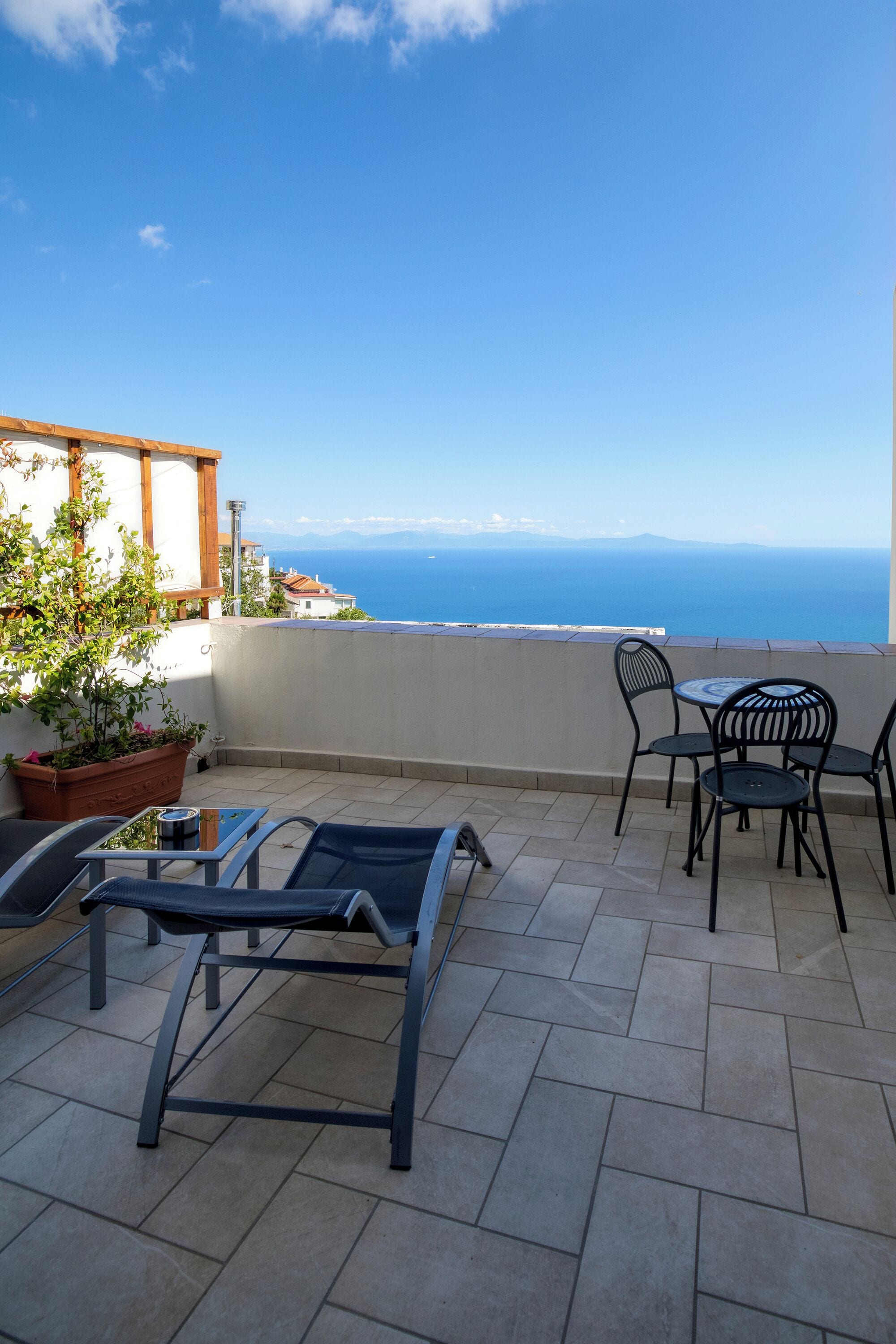 A terrace with a view of the sea, featuring a table and chairs, a potted plant, and a metal chair.