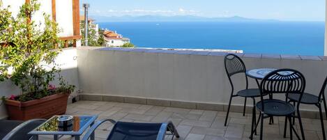 A terrace with a view of the sea, featuring a table and chairs, a potted plant, and a metal chair.
