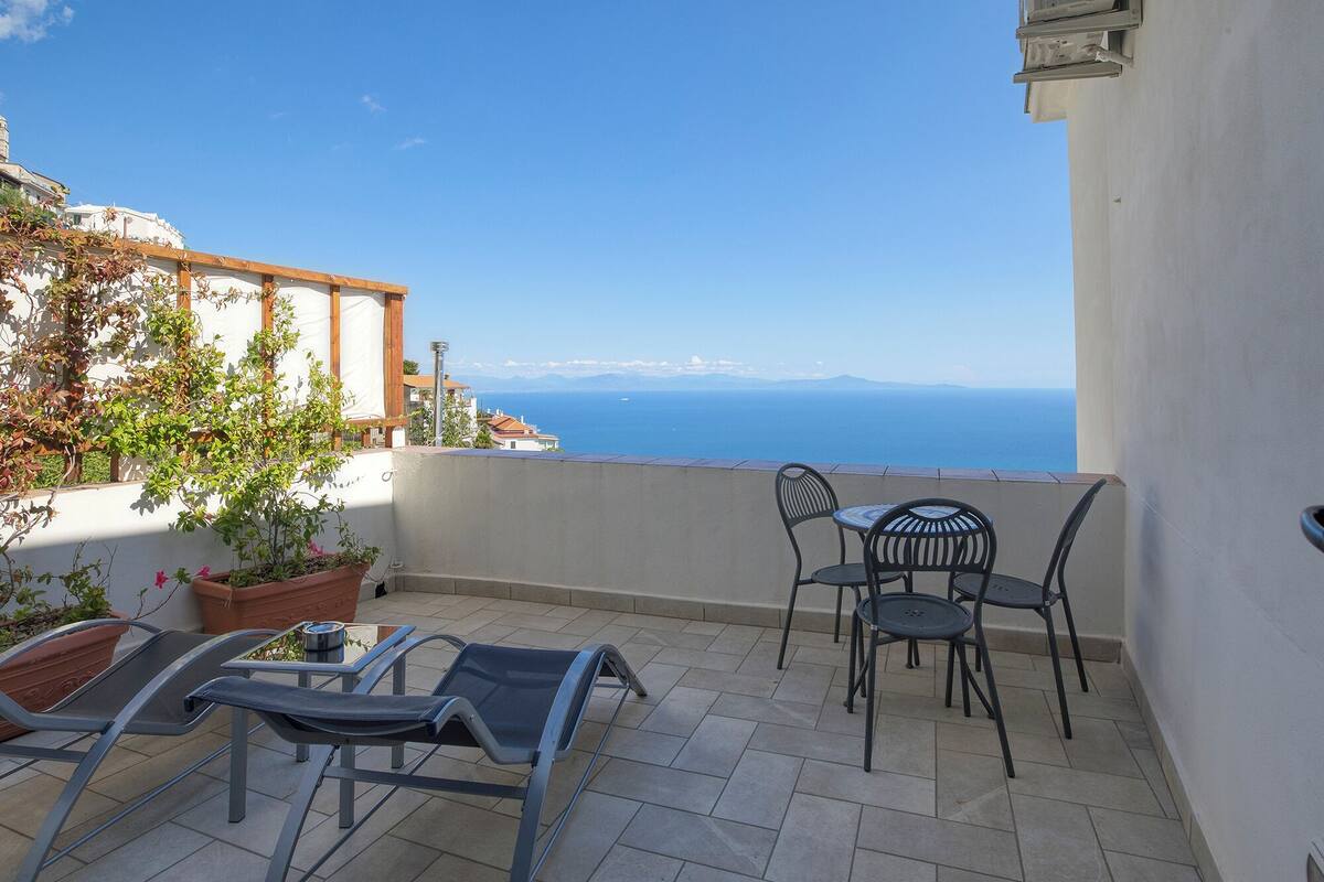 A balcony with a view of the sea, featuring a table and chairs, potted plants, and a tiled floor.