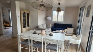 Ein modernes Wohnzimmer mit Essbereich, mit einem weißen Tisch und Stühlen, einem blauen Sofa und einem großen Fenster, das natürliches Licht hereinlässt.