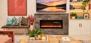 A living room with a fireplace, a sofa with patterned cushions, a coffee table with candles and plants, and a bookshelf with books and decorative items.