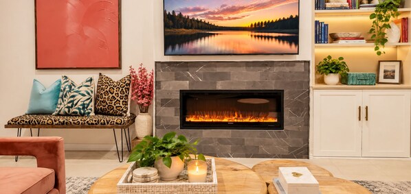 Una sala de estar con chimenea, un sofá con cojines estampados, una mesa de centro con velas y plantas, y una estantería con libros y adornos.