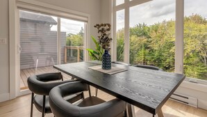 Un comedor con una mesa de madera oscura, sillas negras y un jarrón con flores. Amplias ventanas dejan ver árboles y una casa.