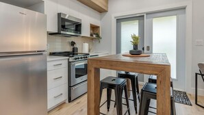 Una cocina moderna con un refrigerador de acero inoxidable, un horno empotrado y un microondas. Hay una mesa de madera con dos taburetes negros y una planta en maceta encima.