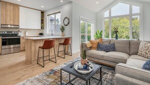 Una cocina moderna con armarios de madera, una isla central y un comedor con dos sillas. Se ve un salón con un sofá y una mesa de centro.