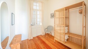 Una habitación con una cama litera de madera, una silla pequeña, un banco de madera, una puerta con cortinas y un cuadro en la pared.