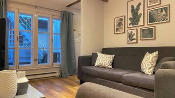 Uma sala de estar com um sofá cinza, almofadas estampadas, uma mesa de centro com objetos decorativos e uma janela com cortinas.