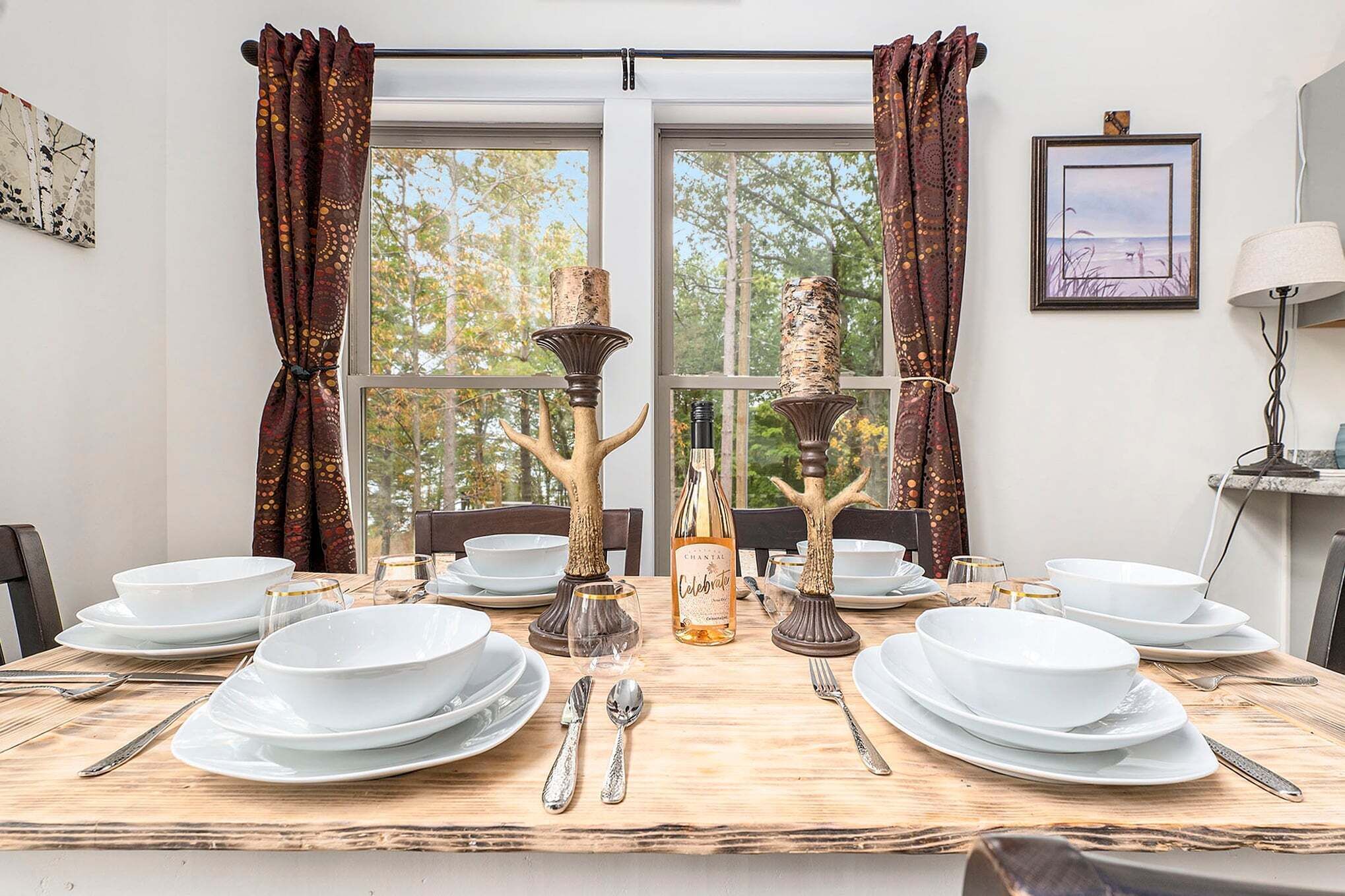 A dining table set with white plates, silverware, and a bottle of champagne, surrounded by windows with curtains.