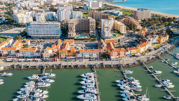 An aerial view of a coastal city with a marina filled with boats, surrounded by residential and commercial buildings. - Apartment Portobello (Loulé)