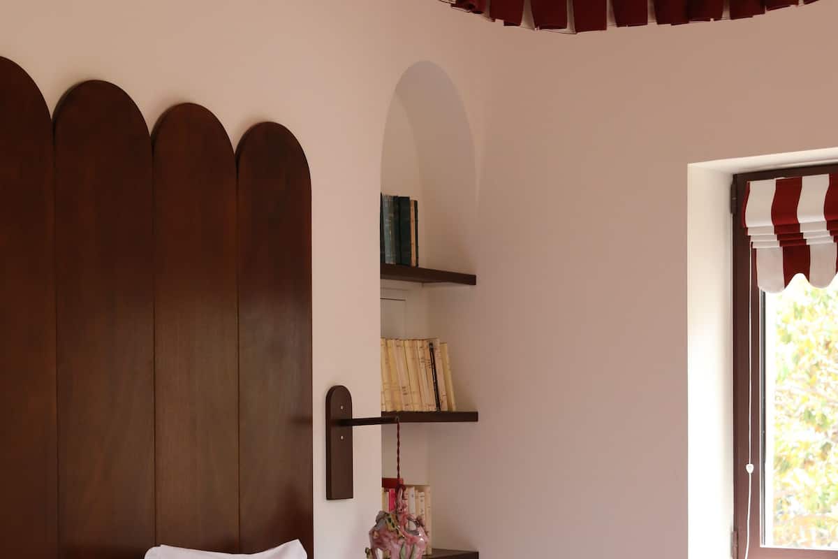 A bedroom with a large bed, a wooden headboard, a red accent wall, a window with striped curtains, and a hanging light fixture.