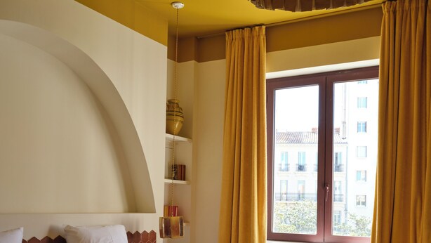 Ein Hotelzimmer mit einem großen Bett, einem Fenster mit Vorhängen, einer Hängelampe und einem Regal mit Büchern und einer Vase.